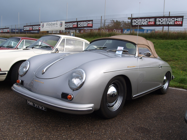 Ein Porsche 356 Speedster auf einem Rasenparkplatz mit anderen Autos, Zäunen, Bannern, Bäumen und einem klaren blauen Himmel.