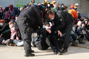 Eine Gruppe von Menschen, einige mit Mützen und Masken, sitzt auf dem Boden neben zwei stehenden Polizeibeamten, mit einer Wand und einer städtischen Umgebung, die auf eine migrantische Festnahmeszene hindeutet.
