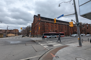 Eine Stadtstraßenszene mit einem Bus an einer Ampel, Fahrzeugen auf der Straße, Passanten auf dem Gehweg, Laternen, Verkehrsampeln, Texttafeln, Bäume, Gebäude mit Fenstern, Strommäste mit Drähten und einem bewölktem Himmel.