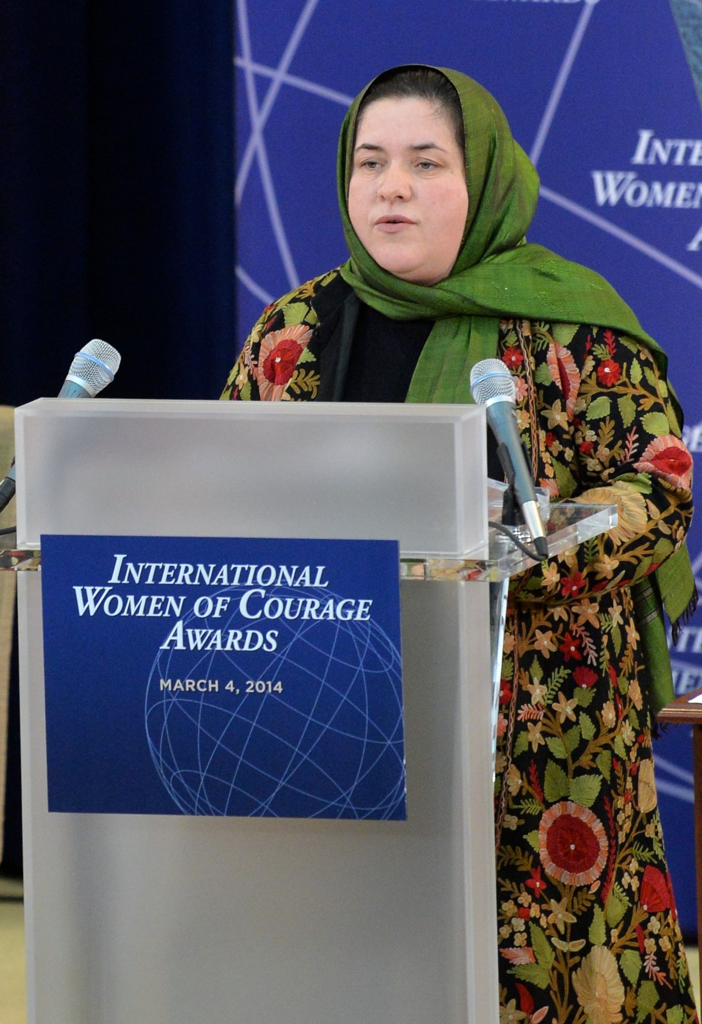 Eine Frau steht an einem Podium mit einem Mikrofon, ein Brett am Podium befestigt und ein Tisch rechts daneben, mit einem Banner im Hintergrund, auf dem 'International Women of Courage Awards 2014' steht.