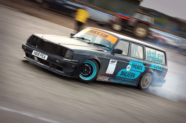 Ein Auto fährt auf einer Rennstrecke, wobei Rauch ausstößt und andere Fahrzeuge sowie Menschen im Hintergrund zu sehen sind.