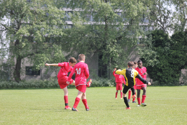 Fünf Jungs in roten T-Shirts und Shorts spielen Fußball auf einem Rasenplatz, mit Bäumen und Gebäuden im Hintergrund.