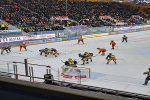 Gruppe von Menschen, die Hockey auf einer Indoor-Eisfläche mit einem Tor spielen, umgeben von einem Zaun, unter Stadionbeleuchtung, mit Zuschauern im Hintergrund und sichtbaren Bannern.