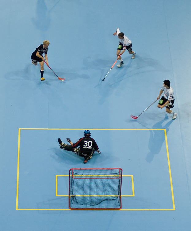 Drei Personen in Sportkleidung spielen Hockey, wobei eine Person rechts mit einem Hockeystock einen Ball schlägt und eine andere Person hinter einem Netz auf dem Boden sitzt.