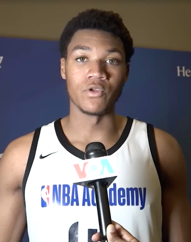 Ein Mann in einem weißen Tanktop hält ein Mikrofon und steht vor einer Tafel, auf der "NBA All-Star Game 2019" steht.