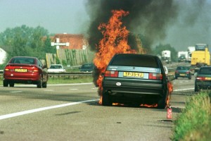 Ein Auto steht in Flammen am Straßenrand, umgeben von anderen Fahrzeugen, mit Bäumen, Gebäuden und einem klaren blauen Himmel im Hintergrund und einem Feuerlöscher auf der rechten Seite neben Gras.