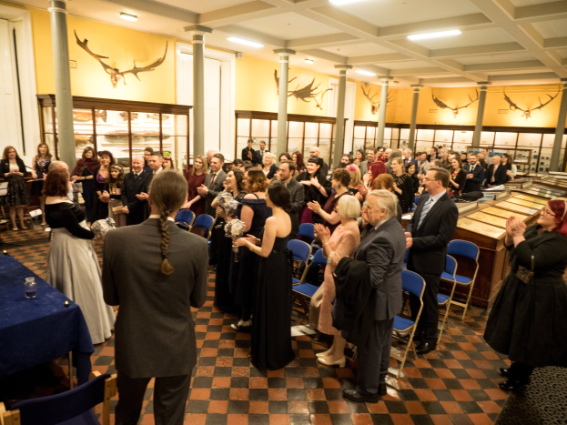 Eine große Gruppe von Menschen steht um einen Tisch in einem Raum mit Stühlen, Tischen, Gläsern, Säulen, Deckenleuchten und Text an den Wänden, der wahrscheinlich auf ein Hochzeitsereignis hinweist.
