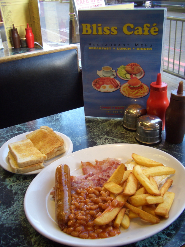 Ein Teller mit Lebensmitteln steht auf einem Tisch mit einer Speisekarte, die 'Bliss Cafe' lautet, Salz- und Pfefferstreuern, Saucen- und Ketchupflaschen und Bannern auf dem Boden, mit einem sichtbaren Fahrzeugrad im Hintergrund.