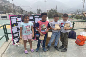 Gruppe von Kindern auf einem Basketballfeld mit Papiertüten in den Händen, mit Gegenständen auf der rechten Seite und einer Tafel mit Text auf der linken Seite, vor einem Hintergrund aus Zaun, Pfosten, Bäumen, Gebäuden, Hügeln und Himmel.