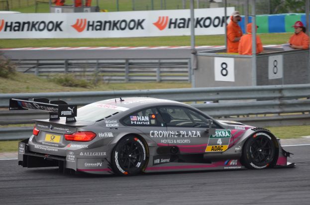 Ein BMW M4 DTM-Rennwagen auf einer Rennstrecke, umgeben von einem Metallzaun, Gras, Pflanzen, einer Gruppe von Menschen, einem Schild, Pfosten, einem Metallrahmen, Bäumen und einem bewölkten Himmel.