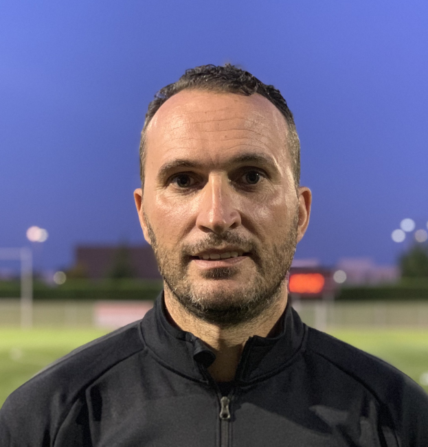 Ein Mann in einer schwarzen Jacke mit Logo steht auf einem Fußballfeld und sieht aus wie ein Trainer mit unscharfem Hintergrund.