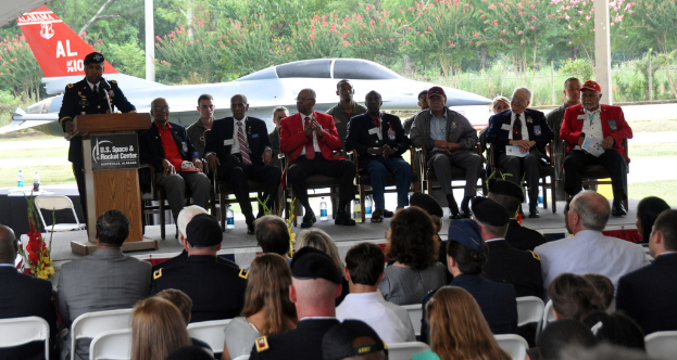 Eine Gruppe von Menschen sitzt im Vordergrund auf Stühlen, im Hintergrund sind auf der B√ľhne einige weitere Personen zu sehen. Eine Person steht links am Rednerpult und spricht in ein Mikrofon mit einem Namensschild davor. Es sind auch Trees, Pflanzen, Pfosten, Wasserflaschen, ein Flugzeug und andere Gegenst√§nde zu sehen.