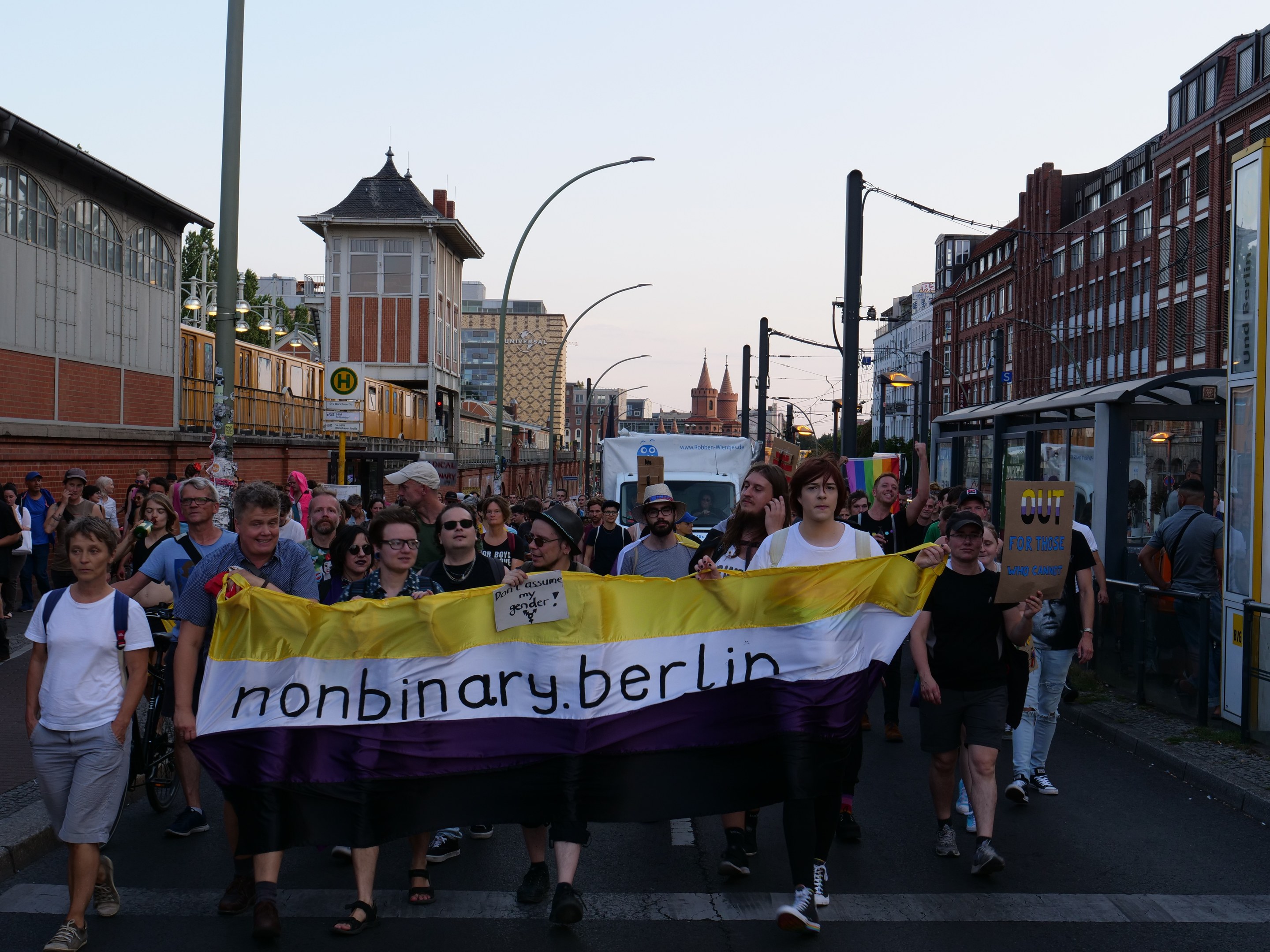Gruppe von Menschen, die eine 'Nonbinary Berlin'-Fahne tragen, auf einer Straße in Berlin mit Laternen, Gebäuden, einer Bushaltestelle, Bäumen und einem klaren blauen Himmel im Hintergrund.