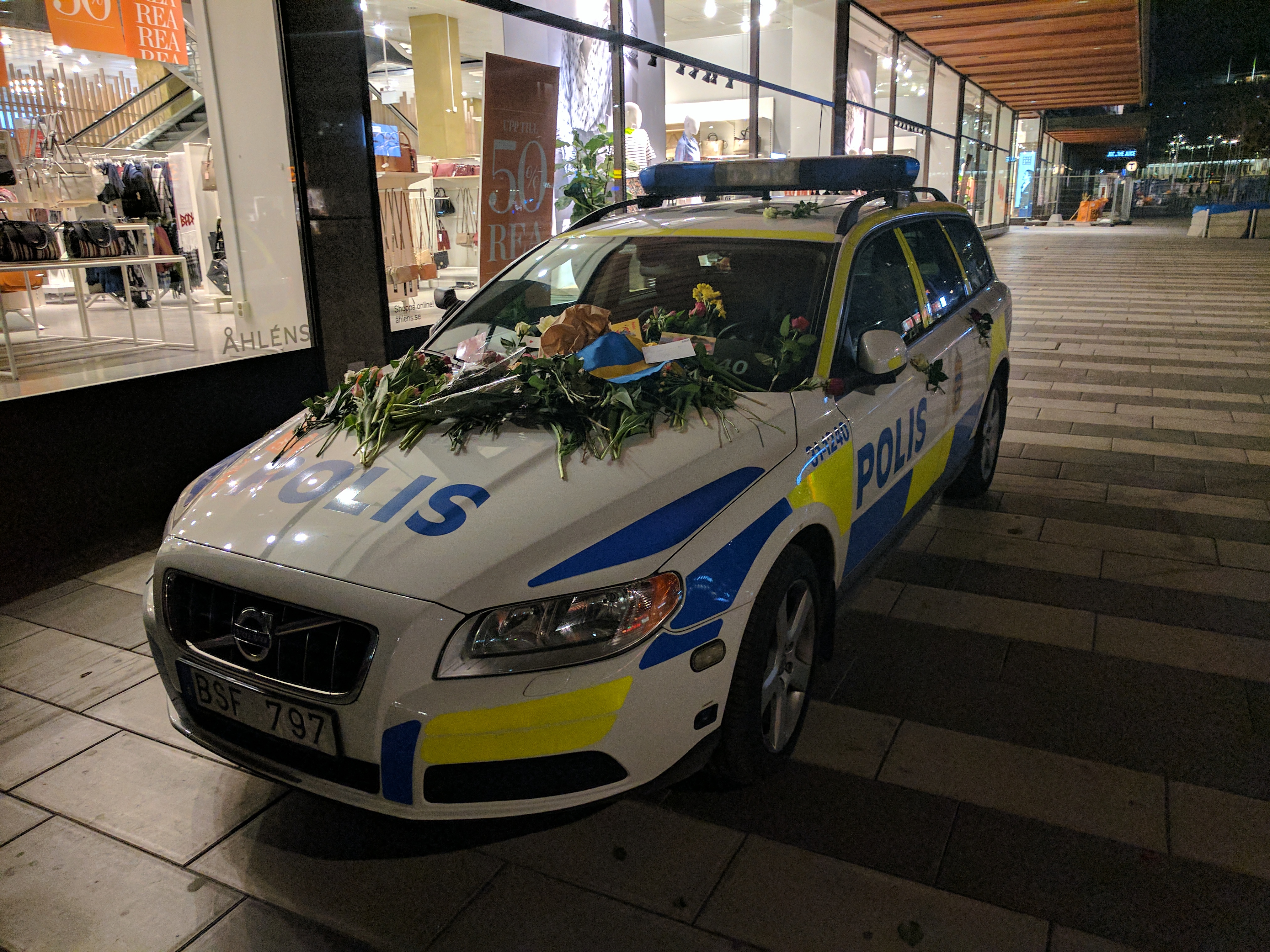 Ein mit Blumen und Blättern geschmücktes Polizeiauto steht nachts vor einem Laden mit Glaswänden, mit Schaufensterpuppen in Kleidern, Texttafeln und anderen Gegenständen im Inneren und Bäumen und Gebäuden im Hintergrund.