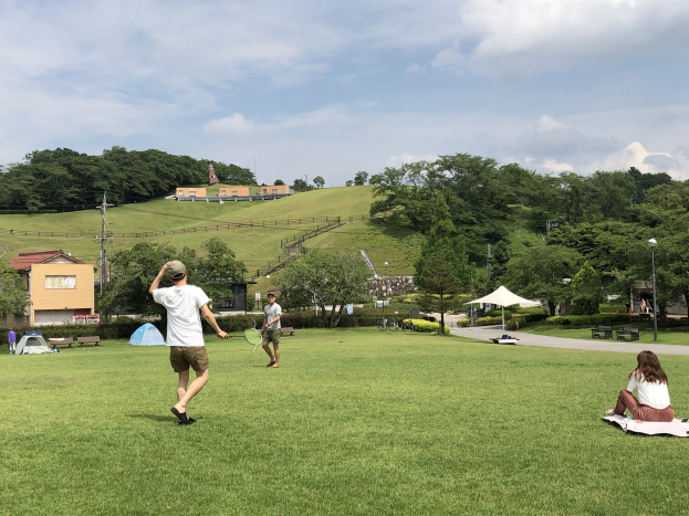 Gruppe von Menschen, die Badminton in einem Park spielt, mit einem Mann, der einen Schläger hält und auf Gras in der Nähe von Zelten und Gebäuden sitzt, mit Hügeln und bewölktem Himmel im Hintergrund.