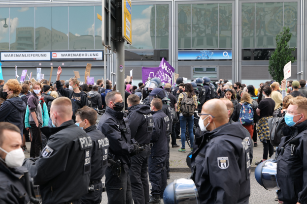 Eine große Gruppe von Menschen steht vor einem Gebäude, einige halten Schilder und tragen Helme, mit einem Hinweisschild im Vordergrund und einem Baum im Hintergrund, was auf eine Protestaktion hindeutet.