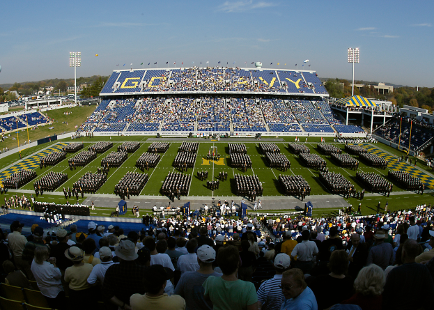 Eine große Menschenmenge in einem Stadion bei einem Fußballspiel, wobei einige stehen und andere sitzen, auf einem grasbewachsenen Feld unter einem klaren blauen Himmel, umgeben von Laternenmasten, Bäumen und Gebäuden.