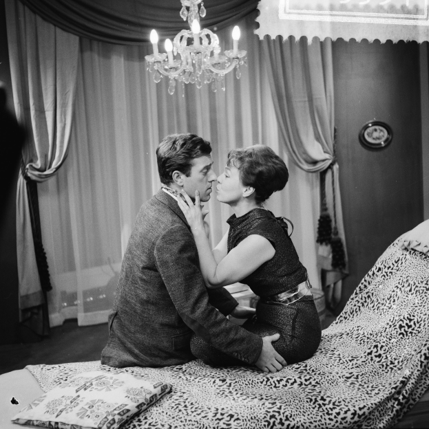 Ein Mann und eine Frau auf einem Bett sitzend, die sich umarmen und küssen, mit Kissen, einer Decke und einem Kronleuchter im Hintergrund und Vorhängen im Raum.