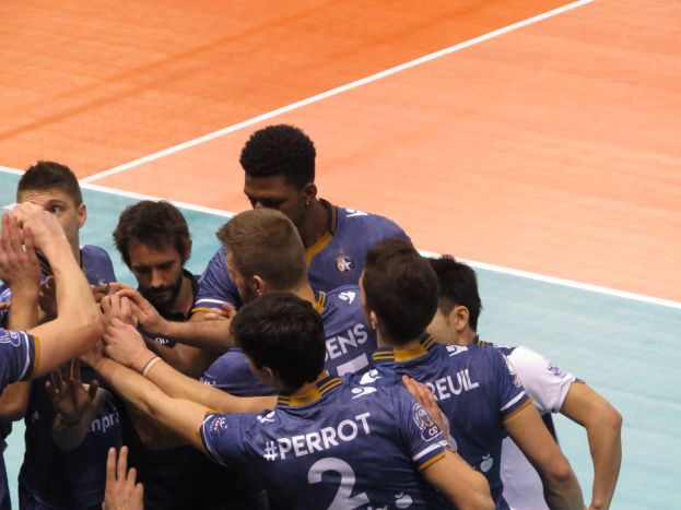 Gruppe von Männern in blauen "Ligue 1"-T-Shirts, die auf einem Volleyballfeld stehen und die Hände siegreich in die Luft recken.