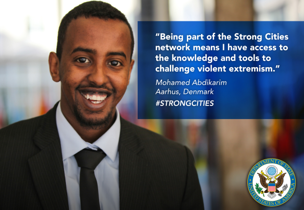 Ein Mann im Anzug und Krawatte lächelt in die Kamera, mit einem Logo und Text auf der rechten Seite, der besagt "Teil des Strong Cities Netzwerks zu sein bedeutet, Zugang zu Wissen und Werkzeugen zu haben, um gewalttätigen Extremismus in Frage zu stellen", und einem unscharfen Hintergrund.