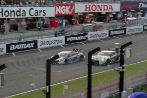 Eine Gruppe von Autos, die auf einer Rennstrecke fahren, umgeben von einem Metallzaun, Gras, Schildern, Metallpfosten, einer Metallabsperrung, einem Zaun, einem Gebäude mit Fenstern und einem Dach mit Deckenleuchten.