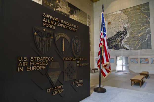 Eingang zum Hauptquartier der US-Strategischen Luftstreitkräfte in Europa mit einer amerikanischen Flagge, Text und Logos an der Wand, Bänken, einer Texttafel, Fotorahmen, beleuchteter Decke und einer Tür im Hintergrund.