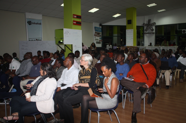 Ein Klassenzimmer mit vielen Menschen auf Stühlen, zwei grüne Säulen mit weißen Tafeln und eine grüne Wand im Hintergrund mit einem Banner und weißen Papieren.