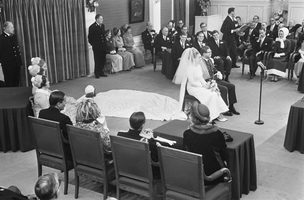 Schwarzes und weißes Foto einer Hochzeitszeremonie mit sitzenden Gästen, einem Mann, der in ein Mikrofon spricht, einer Frau mit einem Blumenstrauß, einem Podium mit Mikrofon, Blumenschmuck, einem Vorhang, einem Rahmen an der Wand und einer Tür im Hintergrund.