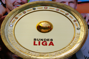 Ein Mann hält ein goldenes Schild mit der Aufschrift "Bundesliga" hoch, im Hintergrund eine Fahne mit drei Personen.