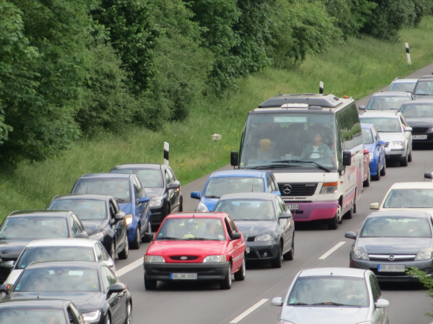 Ein Stau auf einer Autobahn mit vielen Autos und einem Lieferwagen, Menschen sind in den Fahrzeugen zu sehen, und im Hintergrund gibt es Bäume und Gras.