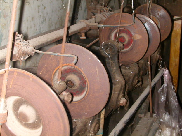 Nahaufnahme einer Metallschleifmaschine mit einem großen zentralen Rad und mehreren kleineren umgebenen Rädern, mit einem Seitengriff, vor einem hellgrauen, schmutzigen Wand.