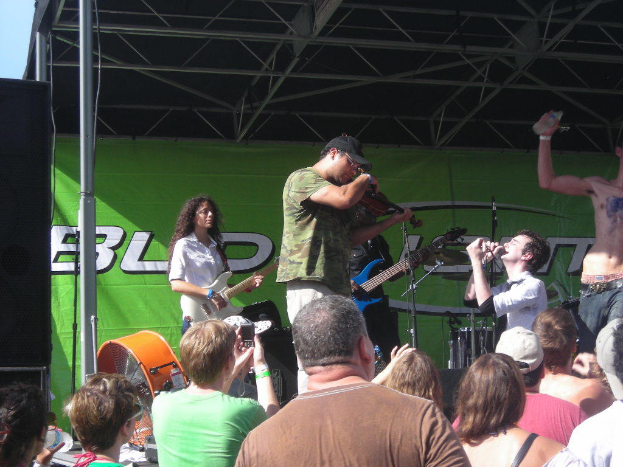 Eine Gruppe von Musikern, die verschiedene Instrumente auf einer Bühne spielen, mit einem großen schwarzen Lautsprecher links daneben, während das Publikum davor steht und die Vorstellung genießt, und ein grünes Banner im Hintergrund.