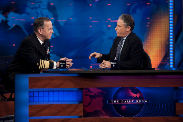 Zwei Manner in schwarzen Blazern, Hemden und Krawatten sitzen an einem Tisch mit Mikrofonen zwischen ihnen und fuhren ein Gesprach vor einem TV-Bildschirm und einem blauen Hintergrund.