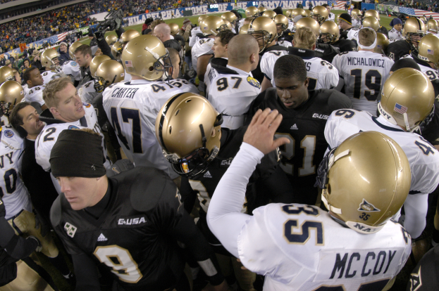 Eine Gruppe von Fussballspielern in Helmen steht zusammen auf einem grünen Feld, mit einer Menge Menschen auf den Tribünen und Transparenten mit Text im Hintergrund.