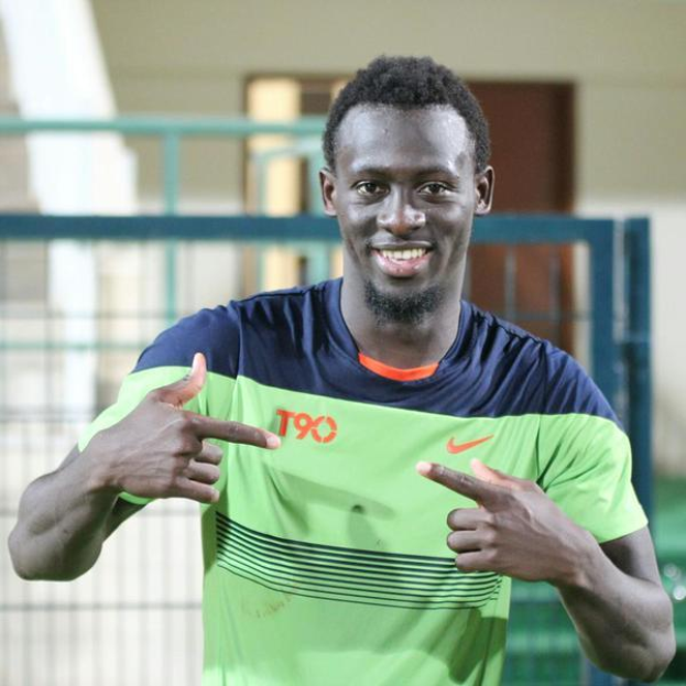 Ein Mann in einem grünen und blauen Hemd, identifiziert als Emmanuel Adebayor, zeigt auf die Kamera und lächelt, mit einem unscharfen Geländer, einer Wand und einer Tür im Hintergrund.