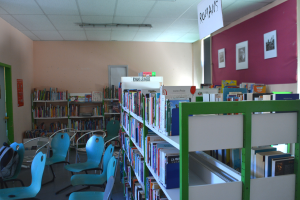 Eine Bibliothek mit Bücherregalen, Stühlen, Taschen auf dem Boden, Wandrahmen, einer Texttafel, einer Tür und Deckenleuchten.