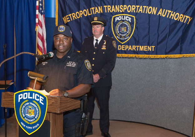 Zwei Polizisten, einer mit Papieren und einem Mikrofon, stehen vor einem Rednerpult, einer Tafel mit Symbolen und Text, einer Fahne, einem Ständer und einem Tisch.