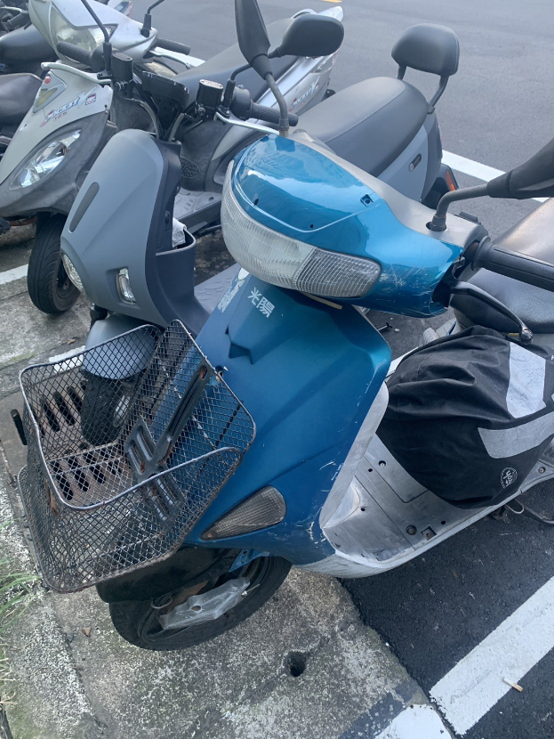 Ein blaues Rollerfahrzeug auf einer Straße neben anderen Motorrädern geparkt, mit Gras links sichtbar.