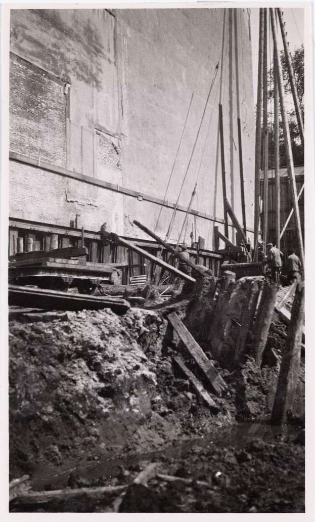 Ein Schwarz-Weiß-Foto einer Baustelle für das New York State Penitentiary, das Arbeiter, hölzerne Gegenstände, Pfähle, Seile, eine Wand und Bäume im Hintergrund zeigt.