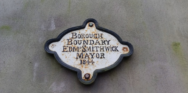 Schwarzes Metallschild mit weißer fettgedruckter Schrift, die 'Borough Boundary Edm Smithwick Mayor' lautet, in der Mitte auf einer Gebäudewand.