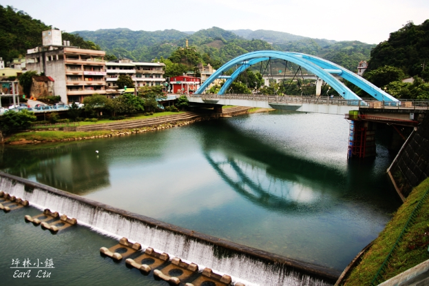 Eine Brücke überspannt einen Fluss mit Gebäuden, Bäumen und Hügeln im Hintergrund bei klarem Himmel.