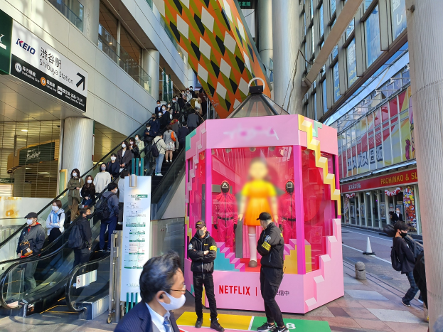 Eine Gruppe von Menschen steht vor einem Gebäude mit Glaswänden, Säulen und Texttafeln, einige tragen Masken, mit einer Rolltreppe im Blick; ein pinker Kasten in der Mitte zeigt Schaufensterpuppen und trägt die Aufschrift "Netflix's new Netflix booth in Tokyo."