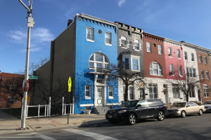 Eine Reihe bunter Häuser an einer Straßenkreuzung in Baltimore, Maryland, mit parkenden Autos, Bäumen, Pfählen, Brettern und einem Zaun, der die Straße säumt, unter einem bewölkten Himmel.