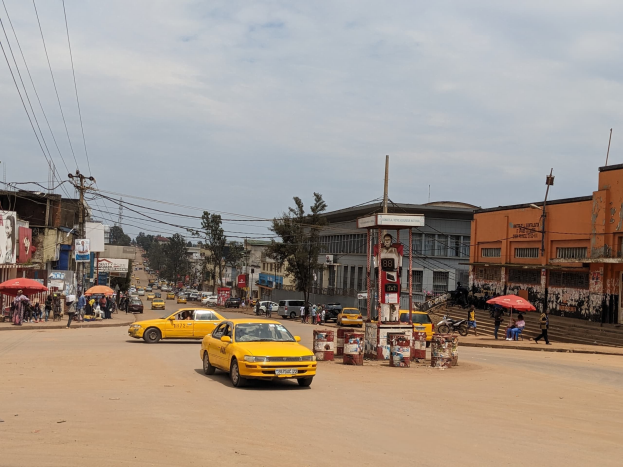 Ein gelbes Taxi fährt auf einer belebten Stadtstraße mit Gebäuden an beiden Seiten, während Fußgänger mit Regenschirmen auf dem Gehweg gehen.