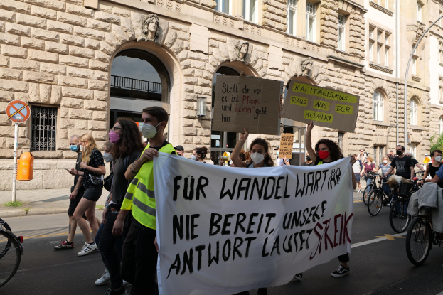 Eine Gruppe von Menschen marschiert auf einer Stadtstraße bei einer Demonstration, einige halten Schilder und andere fahren Fahrräder, im Hintergrund ein historisches Gebäude mit Bögen, Säulen und Skulpturen.