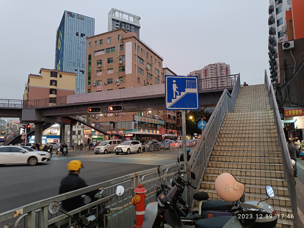 Eine belebte Stadtstraße mit Fahrzeugen, einer Person mit Helm, geparkten Motorrädern, Gebäuden, Verkehrszeichen und einem klaren Himmel.