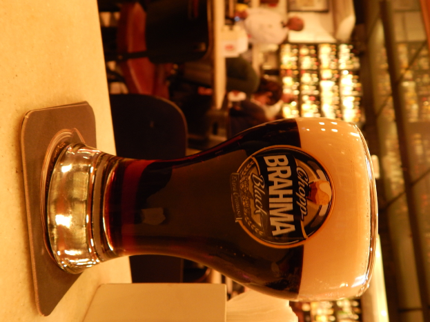 Ein Glas Wein auf einem Teller und eine Papiertaschentuch-Box auf einem Weg, mit einer Gruppe von Menschen, Stühlen, Tischen, einem wandmontierten Rahmen und Lichtern im Hintergrund.