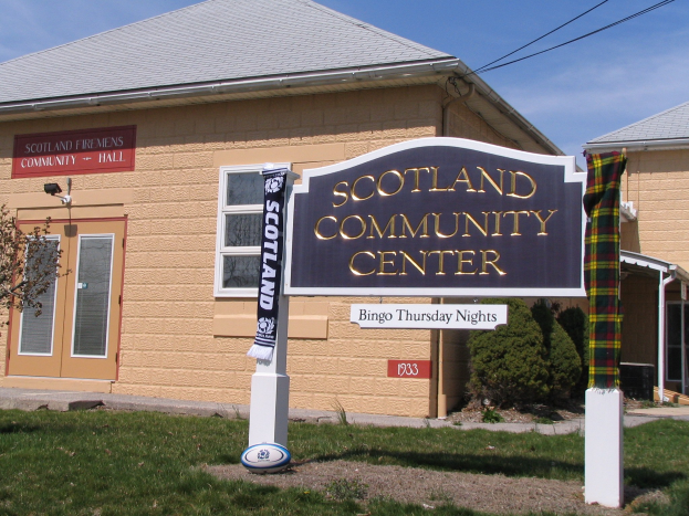Außenansicht des Scotland Community Center mit einem Namensschild, Gras, einem Ball, Pflanzen, einem Baum, sichtbaren Drähten und einem bewölkten Himmel.