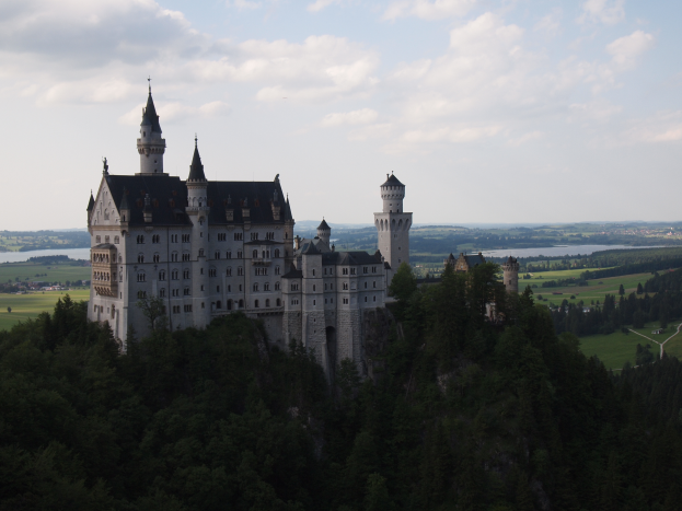 Neuschwanstein Burg in Deutschland, ein majestätisches Gebäude umgeben von saftig grünen Bäumen, einer gewundenen Straße, einem friedlichen Gewässer, sanften Hügeln und einem Himmel voller weißer, flauschiger Wolken.