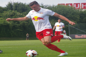 Ein Fussballspieler in Uniform tritt einen Ball auf einem grünen Feld mit Bäumen und einem klaren blauen Himmel im Hintergrund, wobei ein Schild mit der Aufschrift "RB Leipzig" sichtbar ist.